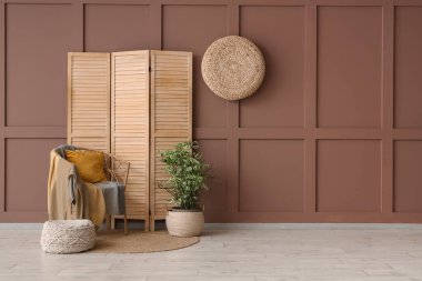 Katlanan ekran, koltuk ve ev bitkisi odanın içindeki kahverengi duvarın yanında.