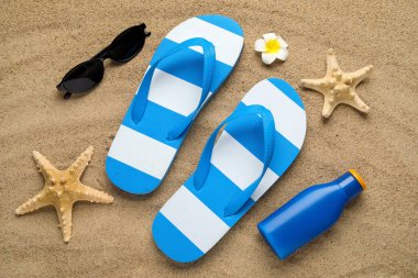 Plaj ayakkabılarıyla kompozisyon, güneş kremi şişesi ve kumun üzerinde güneş gözlüğü, yakın plan.