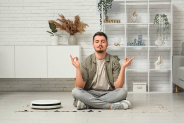 Genç adam evde modern robot elektrik süpürgesiyle yerde meditasyon yapıyor.