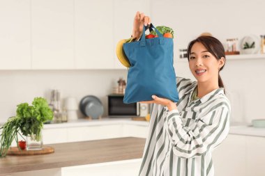 Mutfakta meyve ve sebze dolu mavi alışveriş çantası tutan genç Asyalı kadın.