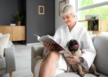 Bornozlu genç kadın evdeki kanepede köpek dergisi okuyor.