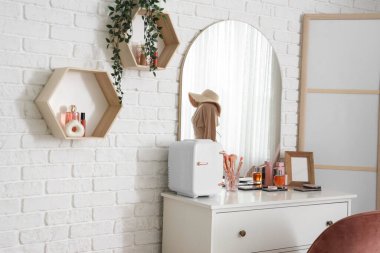 Makyaj masası kozmetik ürünleri, küçük buzdolabı ve hafif tuğla duvarda asılı raflar.