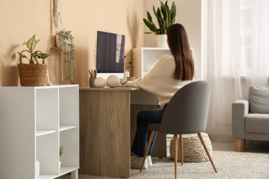 Bej duvarın yanındaki masada modern bilgisayarla çalışan genç bir kadın.