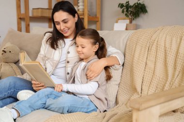 İşitme cihazı olan küçük bir kız ve annesi evdeki kanepede kitap okuyor.