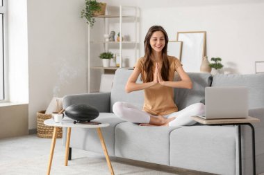 Dizüstü bilgisayarı olan genç bir kadın evdeki kanepede meditasyon yapıyor.