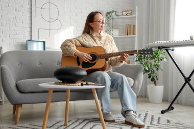 Evdeki kanepede gitar çalan genç bir kadın.