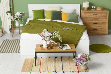 Şık bahar yatak odasının içinde yatak, merdiven, tuvalet ve yeşil duvarın yanındaki masalarda vazolar içinde güzel çiçek buketleri var.