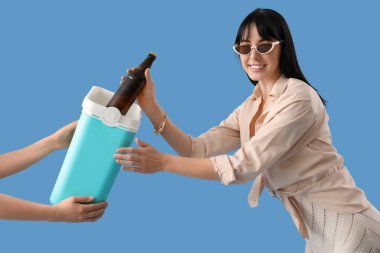 Mavi arka planda taşınabilir buzdolabından soğuk bira alan genç bir kadın.