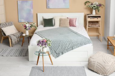 Şık bahar yatak odasının içinde yatak, koltuk, komodin ve güzel çiçek buketleri bej duvarın yanındaki masalarda, üst manzarada.