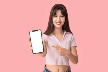 Gülümseyen genç bir kadın pembe arka plandaki boş cep telefonuna işaret ediyor.