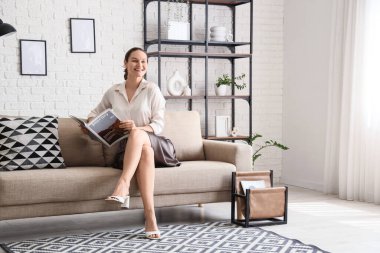 İşten sonra evdeki kanepede dinlenirken dergi okuyan genç bir kadın.