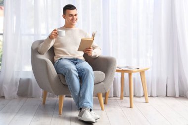 Yakışıklı genç adam kitap okurken kahve içiyor ve pencerenin yanındaki koltukta oturuyor.