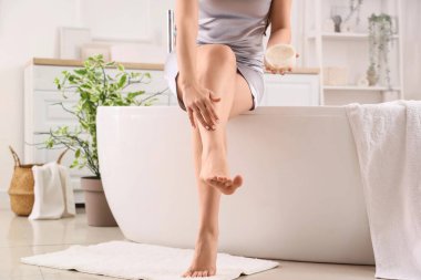 Genç bir kadın banyoda bacaklarına krem sürüyor.