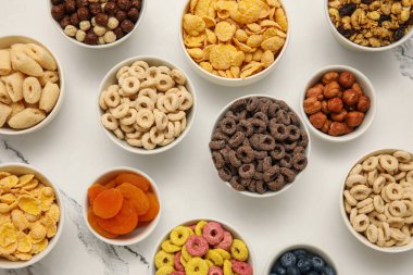 Farklı lezzetli mısır gevrekleri, kuru meyveler ve beyaz arka planda fındıklar.