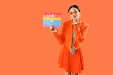 Gökkuşağı bayrağını tutan güzel genç bir kadın LGBT Övünme Ayı ve turuncu arka planda öpüşme