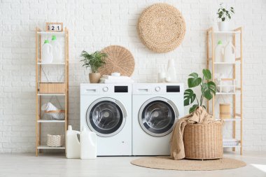 Çamaşır odasındaki beyaz tuğla duvarın yanında çamaşır makinesi, sepet ve deterjan.