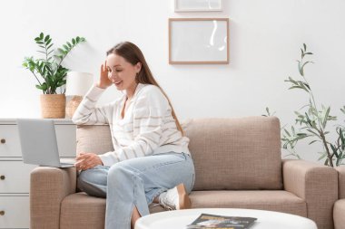 Güzel genç bayan oturma odasındaki şık gri koltukta modern dizüstü bilgisayar kullanıyor.