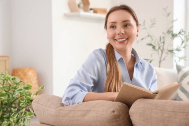Oturma odasındaki gri koltukta kitap okuyan gülümseyen genç kadın.