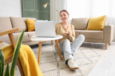 Oturma odasındaki gri kanepenin yanındaki masada dizüstü bilgisayar kullanan gülümseyen genç kadın.