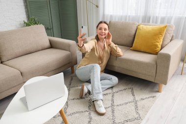Cep telefonuyla gülümseyen genç kadın oturma odasındaki gri kanepenin yanında selfie çekiyor.