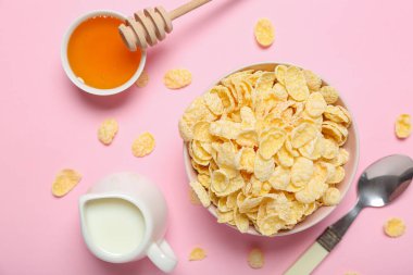 Bir kase lezzetli mısır gevreği, bir sürahi süt ve pembe arka planda bir kase bal içeren kompozisyon, yakın plan.