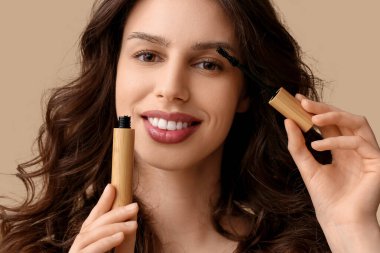 Beautiful young woman applying mascara on brown background