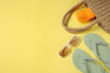 Güneş gözlüklü yeşil parmak arası terlikler, hasır çanta ve sarı arka planda bir şişe güneş kremi.