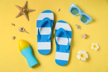 Çiçekli mavi çizgili terlikler, deniz kabukları, bir şişe güneş kremi ve sarı arka planda güneş gözlüğü.