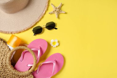 Güneş gözlüklü pembe terlikler, hasır çanta, denizyıldızı, şapka ve sarı arka planda bir şişe güneş kremi.