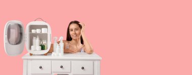 Masanın yanındaki mutlu genç kadın pembe arka planda küçük bir kozmetik buzdolabı açtı.