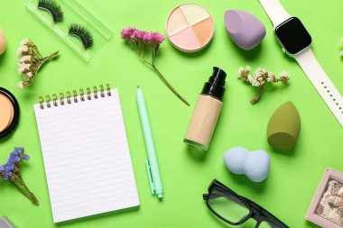 Makyaj malzemesi, modern akıllı saat, gözlük, çiçek ve yeşil arka planda not defteri olan kompozisyon