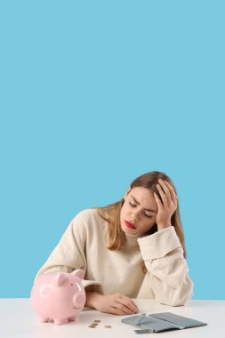 Mavi arka plandaki masada domuz kumbarası, bozuk para ve cüzdanı olan üzgün genç bir kadın. Fiyat artışı kavramı