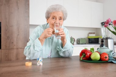 Bir bardak suyla mutfakta hap alan yaşlı bir kadın.