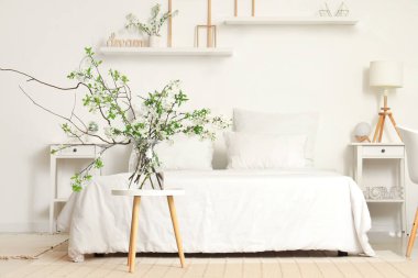 Beyaz yatak odasındaki rahat yatağın yanındaki sehpanın üzerinde çiçek açan bir vazo.