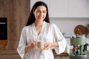 Modern kahve makinesi ve mutfakta bir fincan lattesi olan güzel, genç bir kadın.
