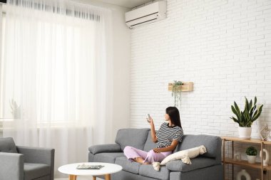 Klimalı uzaktan kumandalı genç bir kadın evdeki beyaz tuğla duvarın yanındaki kanepede oturuyor.