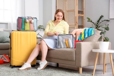 Çantalarında ve bavullarında seyahat aksesuarlarıyla evdeki kanepede oturan hamile bir kadın.