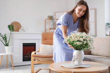 Oturma odasındaki masada papatyaları olan genç bir kadın ve vazo.