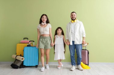 Yeşil duvarın yanında el ele tutuşan bavullu turist ailesi.