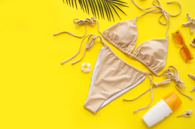 Şık mayolu kompozisyon, güneş kremi şişesi ve sarı arka planda plaj aksesuarları, yakın plan.