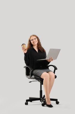 Dizüstü bilgisayarı ve bir fincan kahvesi olan genç iş kadını arka planda sandalyede oturuyor.