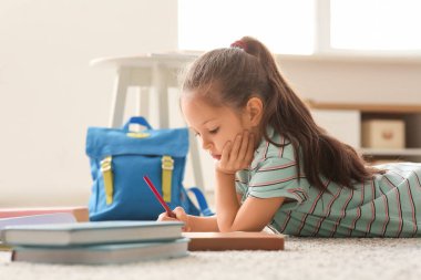 Not defterleri, sırt çantası ve kalemle yerde yatıp evde ders çalışan sevimli küçük kız.