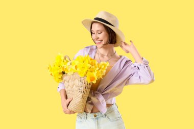 Sarı arka planda bir çanta nergis taşıyan şapkalı genç bir kadın.