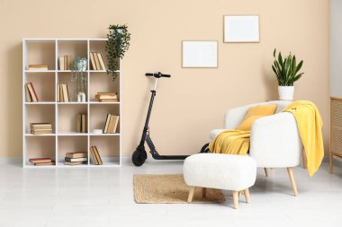 Oturma odasındaki bej duvarın yanında elektrikli scooter, koltuk ve kitaplık.