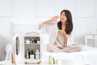 Masada küçük bir kozmetik buzdolabı ve evde yatakta genç bir kadın.