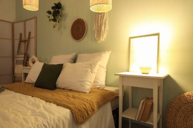 Rahat bir yatak, komodin ve yatak odasının içinde parlayan bir lamba.