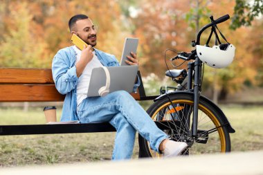 Bilgisayarlı, tabletli ve bisikletli genç mutlu adam parkta bankta cep telefonuyla konuşuyor.