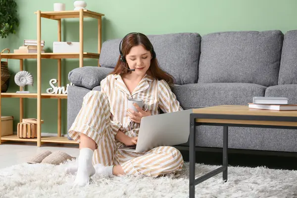 Pijamalı ve kulaklıklı genç bir kadın dizüstü bilgisayarla çalışıyor ve kahve masasının yanında elinde fincanı tutuyor. Uzak çalışma konsepti