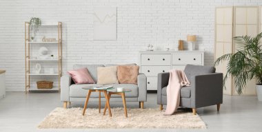 Oturma odasının içinde rahat bir kanepe, koltuk ve beyaz tuğla duvarın yanındaki masalar.