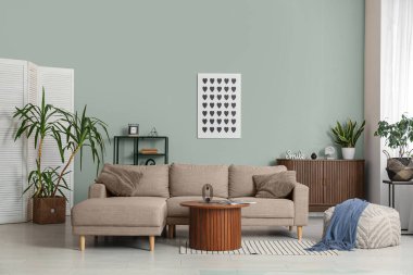 Oturma odasının iç tarafındaki bej duvarın yanında rahat bir kanepe, masa ve puf.